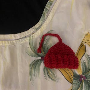 May include: A red crocheted hat with a rounded top and a long, thin, red string attached. The hat is placed on a white fabric with a tropical floral pattern featuring green palm leaves and yellow flowers.