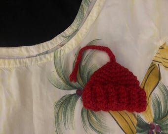 Könnte beinhalten: Ein kleiner, roter Häkelhut mit abgerundeter Oberseite und einem langen, roten Faden. Der Hut befindet sich auf einem weißen Stoff mit einem tropischen Blumenmuster mit grünen Palmenblättern und gelben Blüten.