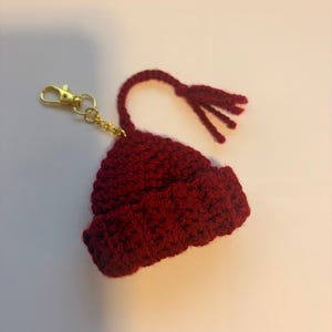 K&ouml;nnte beinhalten: Ein miniaturer, geh&auml;kelter M&uuml;tzen-Schl&uuml;sselanh&auml;nger in tiefem Burgunderrot. Die M&uuml;tze hat einen gerollten Rand und eine kleine Quaste. Ein goldfarbener Verschluss und eine Kette sind zur einfachen Verwendung angebracht.