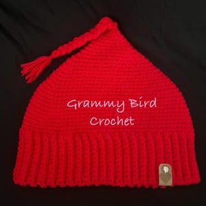 May include: A vibrant red crochet hat with a pointed crown and a plaited tassel. The hat displays the text "Grammy Bird Crochet" and a small tag. The hat is crafted from a soft, textured yarn.