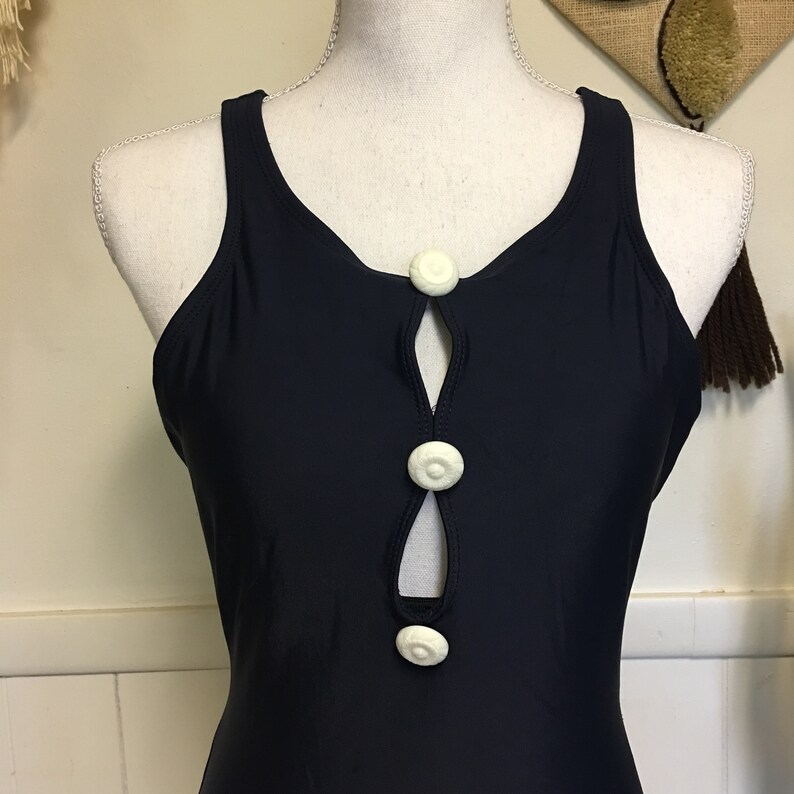 May include: A black swimsuit with a keyhole opening in the front and three white buttons. The swimsuit has a racerback style.