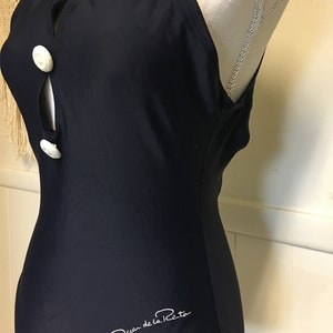 May include: A black one-piece swimsuit with white buttons and the signature "Oscar de la Renta" logo.