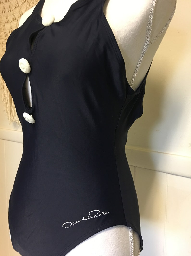 May include: A black one-piece swimsuit with white buttons and the signature "Oscar de la Renta" logo.