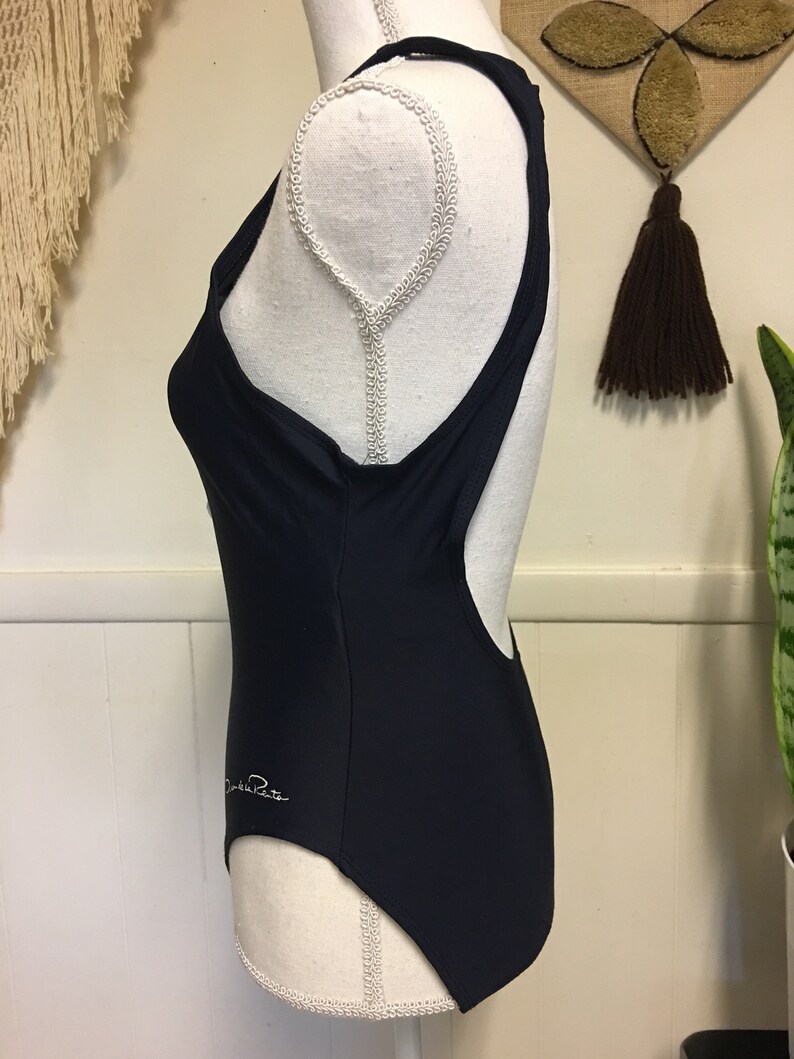 May include: A navy blue one-piece swimsuit with a racerback style and a white "Daniela R" logo on the side.
