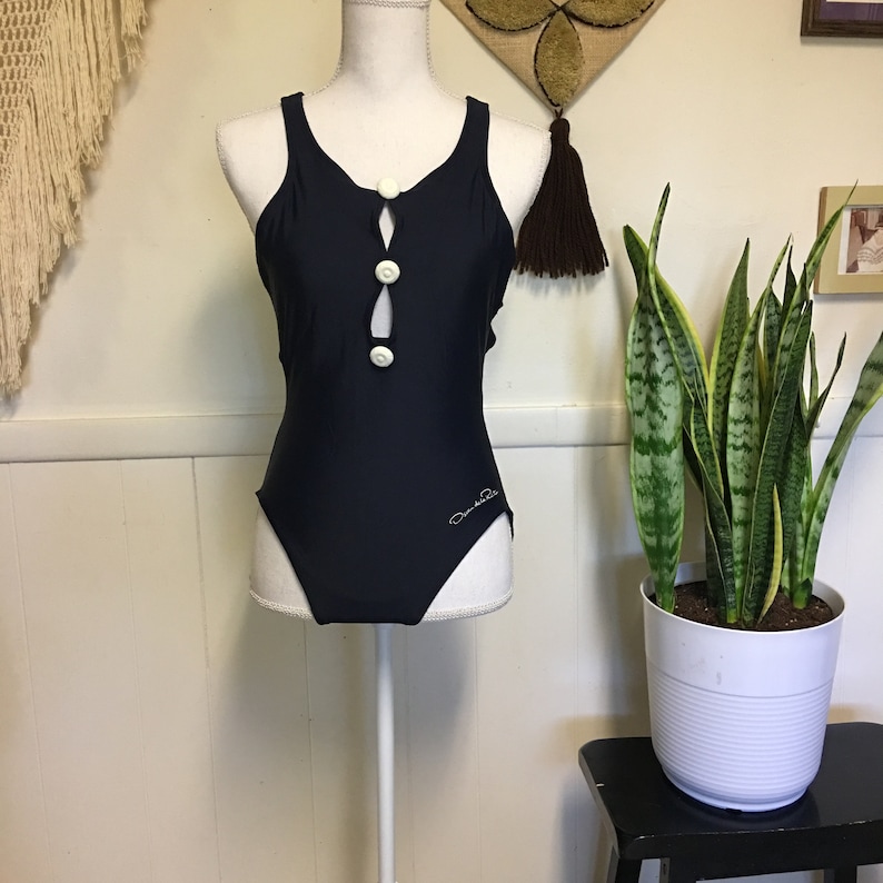 May include: A black one-piece swimsuit with a white button detail down the front. The swimsuit has a scoop neck and a racerback style. The brand name "Daniela" is printed on the bottom right side of the swimsuit.