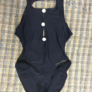 May include: A black one-piece swimsuit with a keyhole opening in the front and three white buttons. The swimsuit has a scoop neck and a racerback style. The brand name "Daniela Retta" is printed on the bottom right side of the swimsuit.