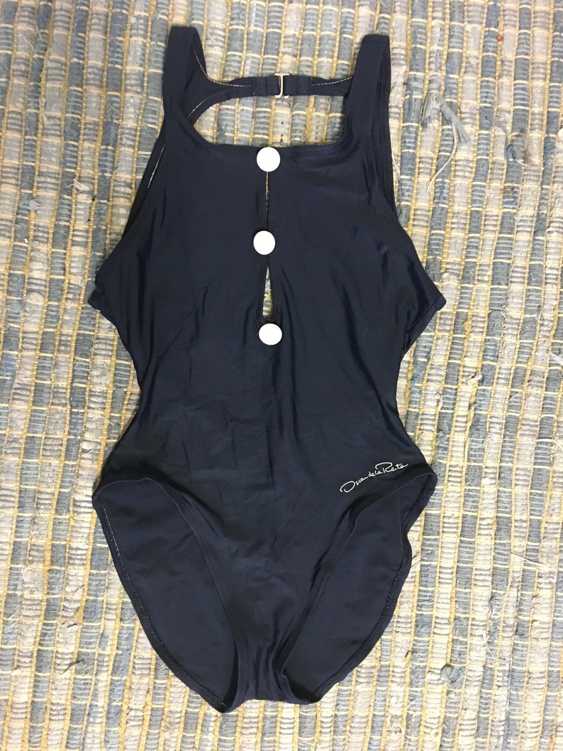 May include: A black one-piece swimsuit with a keyhole opening in the front and three white buttons. The swimsuit has a scoop neck and a racerback style. The brand name "Daniela Retta" is printed on the bottom right side of the swimsuit.