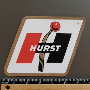 May include: A white, diamond-shaped sticker with a red and black logo. The logo features a stylized shifter with a red ball on top and the word "HURST" in black letters.