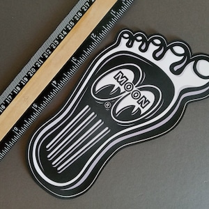 May include: Black and white sticker of a footprint with the word "MOON" in a stylized font. The footprint has a white outline and the word "MOON" is in a white circle.