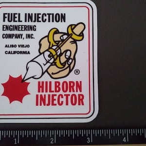 Könnte beinhalten: Quadratischer Aufkleber mit weißem Hintergrund und rotem und schwarzem Rand. Der Aufkleber zeigt den Text "FUEL INJECTION ENGINEERING COMPANY, INC." und "HILBORN INJECTOR". Eine Cartoon-Hand hält eine Spritze. Der Aufkleber ist ca. 7.6 cm breit.