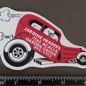 May include: A red vintage car with a white background. The car has a white driver in the driver's seat. The car has the text "JARDINE HEADERS 7565 ACACIA GARDEN GROVE 893-0913" on the side.