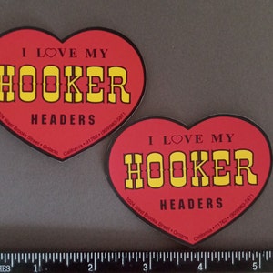 Puede incluir: Dos pegatinas en forma de corazón con el texto "I LOVE MY HOOKER HEADERS" en amarillo y negro sobre un fondo rojo. Las pegatinas también incluyen la dirección y la información de contacto. Una regla en pulgadas en la parte inferior.