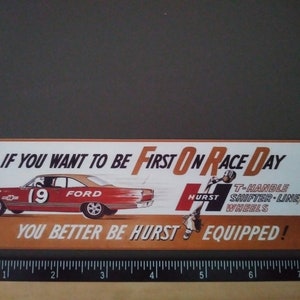 May include: Vintage rectangular sign with a red Ford race car, the number 19, and the text "IF YOU WANT TO BE FIRST ON RACE DAY YOU BETTER BE HURST EQUIPPED!" The sign also features the Hurst logo.