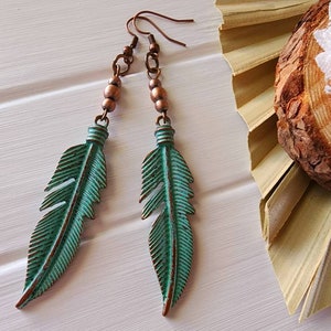 Puede incluir: Un par de aretes largos de plumas azul turquesa con cuentas de cobre y alambre. Los aretes cuelgan de una superficie blanca.