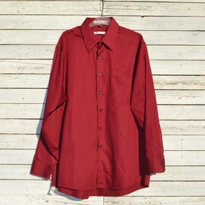 Puede incluir: Una camisa de manga larga con botones, de color rojo intenso, colgada frente a una pared de madera pintada de blanco desgastado. La camisa presenta un cuello clásico, un bolsillo en el pecho y puños abotonados. La prenda se muestra en una percha.