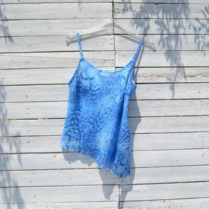 May include: A blue sleeveless top with a white background and a blue abstract pattern. The top is hanging on a white wooden hanger against a white wooden background.