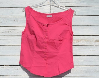 May include: A sleeveless pink button-up top with a rounded hemline. The top is hanging on a white wooden background.