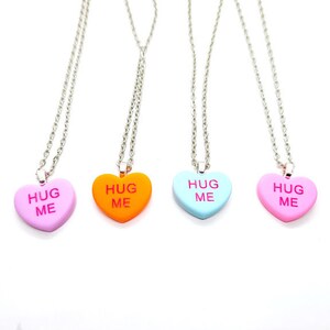 Op de afbeelding: Vier zilveren kettingen met hartvormige hangers. Elke hanger is in een andere kleur: roze, oranje, blauw en lichtblauw. De hangers hebben allemaal de woorden "Hug Me" in roze erop geschreven.