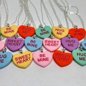 Op de afbeelding: Een set van 12 zilveren kettingen met hartvormige hangers. De hangers zijn in verschillende kleuren en hebben verschillende berichten, waaronder "Sweet Heart", "Hug Me" en "Be Mine".
