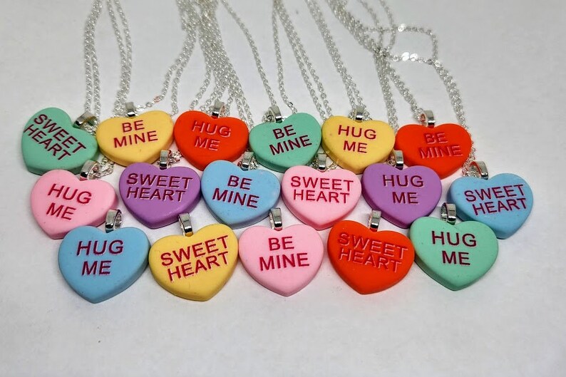 Op de afbeelding: Een set van 12 zilveren kettingen met hartvormige hangers. De hangers zijn in verschillende kleuren en hebben verschillende berichten, waaronder "Sweet Heart", "Hug Me" en "Be Mine".