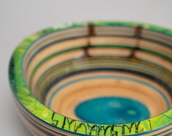 Può includere: Una ciotola di legno realizzata con skateboard riciclati. La ciotola è composta da strati di legno di diversi colori, tra cui verde, blu e marrone. L'interno della ciotola è dipinto di un blu brillante.