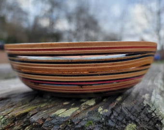 Può includere: Una ciotola di legno realizzata con skateboard riciclati. La ciotola presenta un design a strati con colori alternati di marrone, nero, rosso e blu.