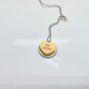 Op de afbeelding: Een zilveren ketting met een gele hartvormige hanger met de tekst "BE MINE" in rode letters.
