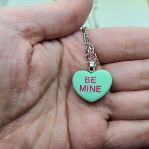 Op de afbeelding: Een zilveren ketting met een groene hartvormige hanger waarop in roze letters "BE MINE" staat.
