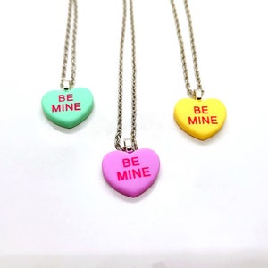 Op de afbeelding: Drie zilveren kettingen met hartvormige hangers. Elke hanger is in een andere kleur: groen, roze en geel. De hangers hebben allemaal de woorden "BE MINE" in rood.