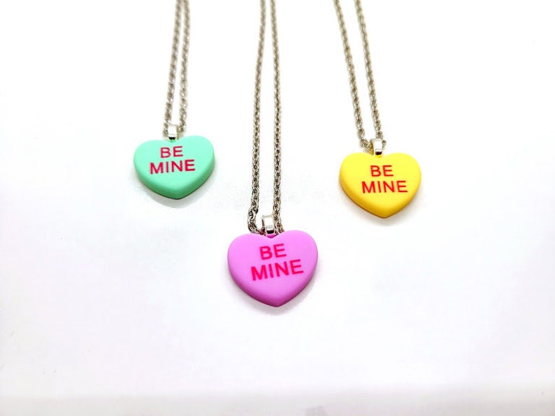 Op de afbeelding: Drie zilveren kettingen met hartvormige hangers. Elke hanger is in een andere kleur: groen, roze en geel. De hangers hebben allemaal de woorden "BE MINE" in rood.