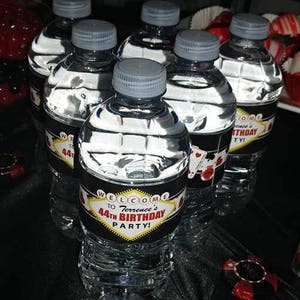 May include: A set of six clear plastic water bottles with black and red labels. The labels have a Las Vegas theme and say "Welcome to Terrence's 44th Birthday Party!"