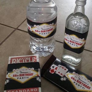 May include: A set of party favors for a 44th birthday party. The favors include a bottle of water, a bottle of liquor, a chocolate bar, and a deck of cards. All of the items have labels with a Las Vegas theme and the text "Welcome to Terrence's 44th Birthday Party!"