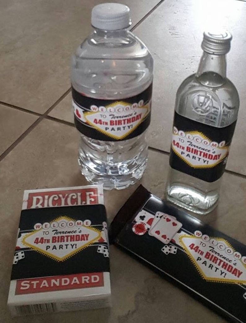 May include: A set of party favors for a 44th birthday party. The favors include a bottle of water, a bottle of liquor, a chocolate bar, and a deck of cards. All of the items have labels with a Las Vegas theme and the text "Welcome to Terrence's 44th Birthday Party!"