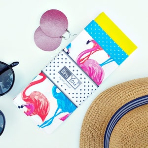 Könnte beinhalten: Ein weißes Geschirrtuch mit einem blauen und rosa Flamingo-Druck. Das Geschirrtuch hat einen schwarz-weißen Polka-Dot-Rand und die Worte "Bless You" in einer schwarz-weißen Schrift. Das Geschirrtuch ist in der Hälfte gefaltet und liegt auf einer weißen Oberfläche.