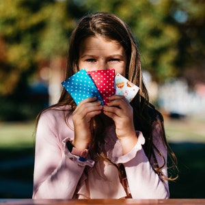 Könnte beinhalten: Ein junges Mädchen hält drei Stoffgeschenktüten vor seinem Gesicht. Die Taschen sind blau mit weißen Punkten, rot mit weißen Punkten und weiß mit einem bunten Cupcake-Design.