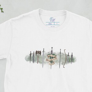 Op de afbeelding: Wit T-shirt met een aquarel-illustratie van zwaarden en bijlen. De zwaarden zijn in een rij gerangschikt en hebben verschillende ontwerpen. De tekst "Winter is Coming" staat op een schild in het midden van de illustratie.