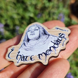 May include: A wooden pin featuring a black ink illustration of a person with a head covering, looking upward with a neutral expression. The text "I hate mondays!" is written in a stylized, dark script font across the bottom of the wooden surface.