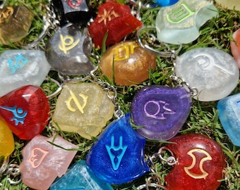May include: A collection of colorful resin keychains with various symbols and designs. The keychains are in shades of blue, red, yellow, green, and purple, with silver key rings. The keychains are scattered on a bed of green grass.