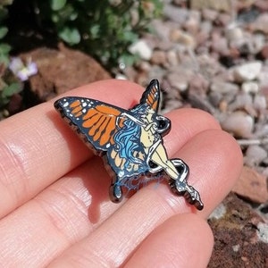 May include: A black metal pin with a colorful butterfly design. The butterfly has orange, yellow, and blue wings with a white and blue fairy figure inside.