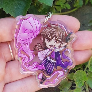 May include: A clear acrylic charm featuring a manga-style character with brown hair, a purple outfit, and a determined expression. The charm includes a silver-colored keychain and a pink energy effect. The background shows green foliage.