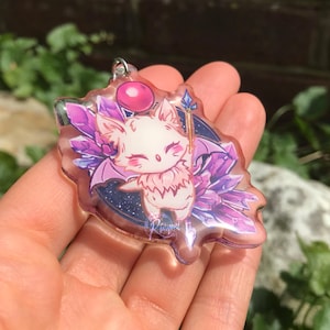 May include: A clear acrylic keychain featuring a cartoon character with a pink and white color scheme. The character has wings, a staff, and is surrounded by purple crystals and a pink balloon. The keychain has a silver metal ring.