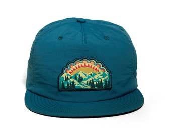 May include: A teal blue baseball cap with a colorful embroidered patch of a mountain scene with a sunrise. The patch features a yellow sun with red and orange rays, green trees, and white snow-capped mountains.