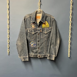 May include: A vintage denim jacket with multiple patches. The jacket is a medium wash blue with a classic button-up design. Patches include the word "HAWK" and other circular designs. The jacket is hanging on a hanger.