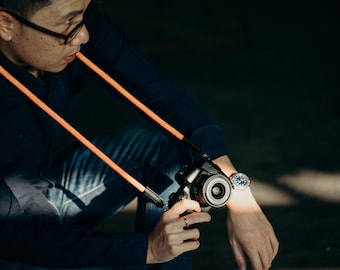 Könnte beinhalten: Ein leuchtend orangefarbener Seil-Kameragurt mit schwarzen Lederenden, der an einer schwarzen Digitalkamera befestigt ist. Der Gurt wird um den Hals getragen und verfügt über ein strukturiertes, geflochtenes Design, das einen Kontrast bildet.