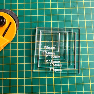 Op de afbeelding: Een transparante acryl liniaal met vijf verschillende vierkanten die in inches zijn gemarkeerd. De vierkanten zijn geëtiketteerd met 1 vierkant, 1" vierkant, 1 1/2 vierkant, 1 1/2" vierkant en 2" vierkant.