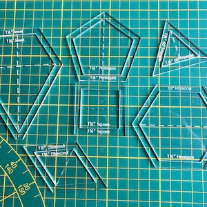 May include: A set of clear plastic quilting templates for creating various shapes, including a 1 1/2 inch hexagon, a 1 1/2 inch pentagon, a 1 1/2 inch square, a 1 1/2 inch diamond, a 1 1/2 inch jewel, and a 1 1/4 inch 60 degree triangle. The templates are laid out on a green cutting mat.
