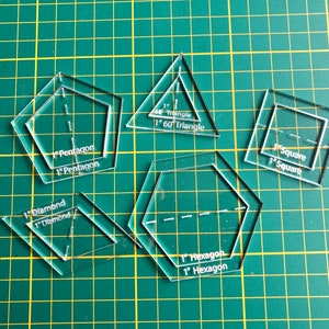 Op de afbeelding: Transparante acryl quilt sjablonen in verschillende geometrische vormen: vijfhoek, driehoek, vierkant, ruit en zeshoek. Elke sjabloon is gemarkeerd met zijn vorm en grootte, 2,54 cm. De sjablonen zijn gerangschikt op een groene snijmat.