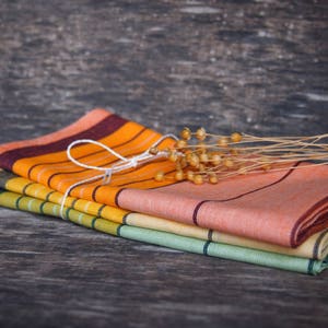 May include: A stack of four striped kitchen towels in orange, yellow, green, and brown colors. The towels are folded and tied together with a white string. The towels are on a wooden surface.