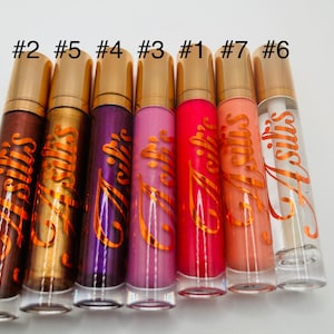 May include: Seven clear lip gloss tubes with gold lids and labels that say "Asillis". The lip glosses are in various colors, including brown, gold, purple, pink, red, orange, and clear.