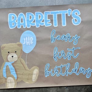 K&ouml;nnte beinhalten: Braunes Papierschild mit blauem Text: "Barrett's beary first birthday". Ein Teddyb&auml;r mit blauem Band und ein blauer Ballon mit den Initialen "JHB" sind ebenfalls abgebildet. Die R&auml;nder haben ein blau-wei&szlig;es Karomuster.